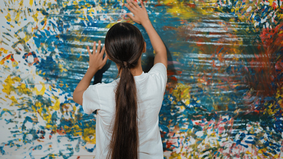 Back view of young cute girl raise hand to paint the colorful stained wall. Attractive highschool student paint the wall with hand print while holding color palette. Creative activity. Edification.