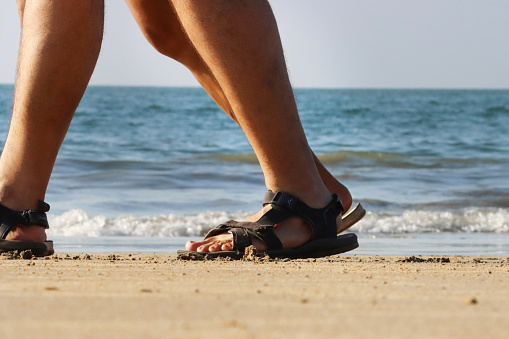 Stock photo of unrecognisable people wearing sandals walking along sunny, golden sandy beach with sea at low tide in the background.