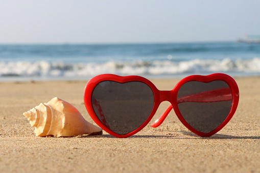 Stock photo showing close-up view of red, heart-shaped sunglasses besides a helix seashell on a sunny, golden sandy beach with sea at low tide in the background.