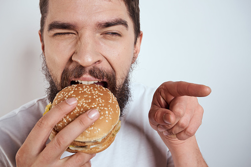 A man eating a hamburger on a light background in a white T-shirt cropped view close-up hunger fast food. High quality photo