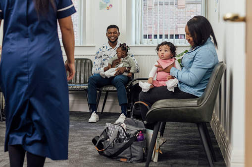 A shot of a small group of parents sitting together with their babies in the waiting room of a baby clinic in Newcastle-upon-Tyne, North East England. One of the babies is looking up at the unrecognisable nurse who is walking towards them, whilst her mother is in side profile view. A father with baby is smiling behind.