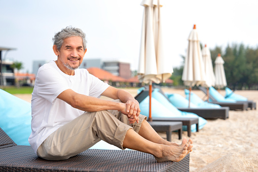 portrait mature male sitting on beach chair,feeling freedom,asian senior man having relaxation at seaside on summer time,concept of elderly pensioner lifestyle,holiday,travel,wellness,wellbeing