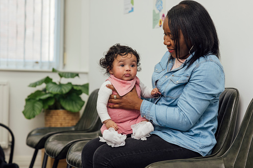 A shot of a mother and her baby in the waiting room of a baby clinic in Newcastle-upon-Tyne, North East England. The baby is sitting up on her mother's knee whilst she winds her, one hand supporting her front. The baby looks ahead, off-frame, as her mother smiles down at her.