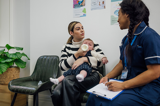 A shot of a young mother sitting with her baby and a nurse in the waiting room of a baby clinic in Newcastle-upon-Tyne, North East England. She is feeling her baby with a bottle whilst talking to the nurse who holds a clipboard containing paper documents.