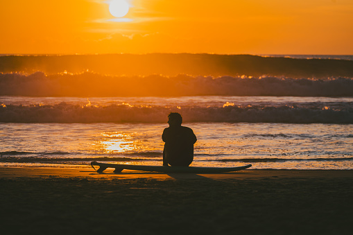 Silhouette of unrecognizable surfer sit on the sand along the shoreline, with waves crashing in the background, during a breathtaking sunset