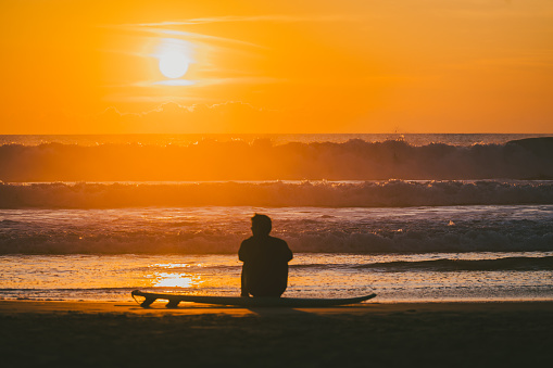 Silhouette of unrecognizable surfer sit on the sand along the shoreline, with waves crashing in the background, during a breathtaking sunset