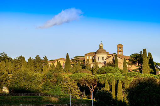 A beautiful cityscape of the Colle Celio (Caelian Hill) in Rome seen from the gardens of the Aventine hill, in the heart of the Eternal City. On the horizon the ancient Basilica dei Santi Giovanni e Paolo (Basilica of Saints John and Paul) built starting from 398 AC, and in the foreground the gardens of San Gregorio. The Caelian Hill, one of the seven ancient hills of Rome, between the Aventine and the Esquiline hills, preserves numerous archaeological areas from the republican and imperial era, such as the Temple of Divus Claudius, and important churches such as the Basilica of Santi Quattro Coronati and the Basilica of Saints John and Paul. In 1980 the historic center of Rome was declared a World Heritage Site by Unesco. Image in high definition quality.