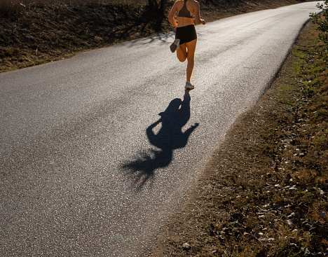 Shadow cast on a paved road of a fit young female runner running with a perfect style and her hair loose in the air on a paved mountain trail with the dawn light backlit creating glare on the ground.