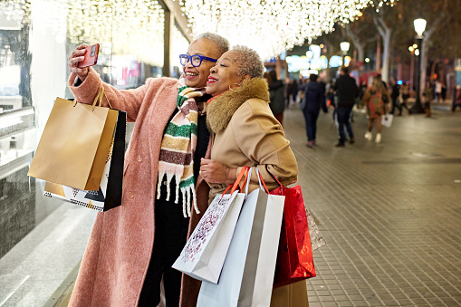 Well dressed Black women in 50s and 60s wearing warm clothing, carrying bags, standing beneath glittering lights and capturing a memory with smart phone.
