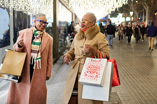 Women in 50s and 60s dressed for winter, carrying bags, talking and laughing as they walk outdoors beneath glittering decorations.