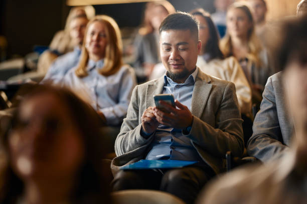 uśmiechnięty chiński biznesmen korzystający z telefonu komórkowego na seminarium w centrum kongresowym. - chmara zdjęcia i obrazy z banku zdjęć