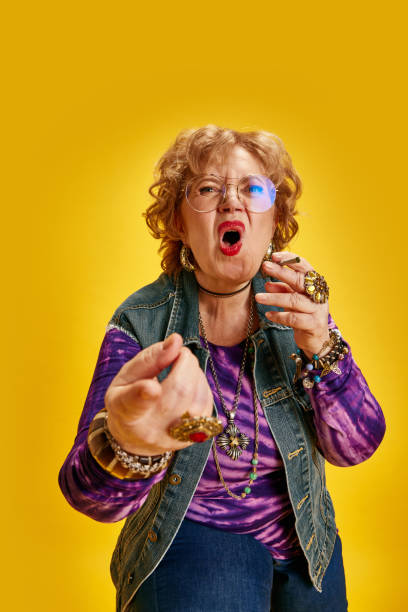 istock Vocal elderly woman gesturing forward looking ta camera dressed denim jacket purple shirt and ornate accessories against yellow background. 1962218405