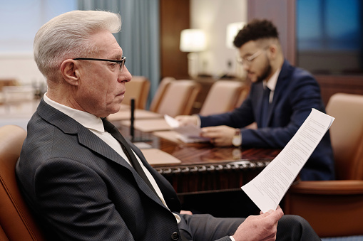 Mature businessman reading documents in boardroom, his young colleague working on background