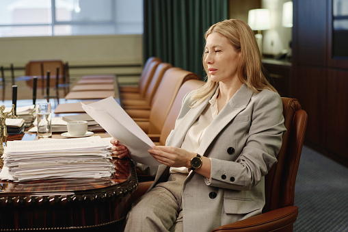 Experienced business woman reading document from pile of papers placed on table in conference room