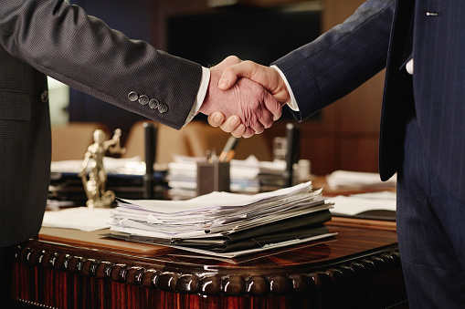 Closeup of two unrecognizable businessmen shaking hands standing next to table with pile of papers