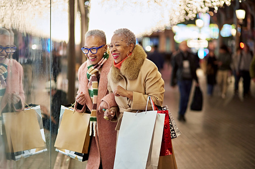 Well dressed black women in 50s and 60s carrying bags and laughing as they pause beneath glittering lights to admire merchandise in retail window.