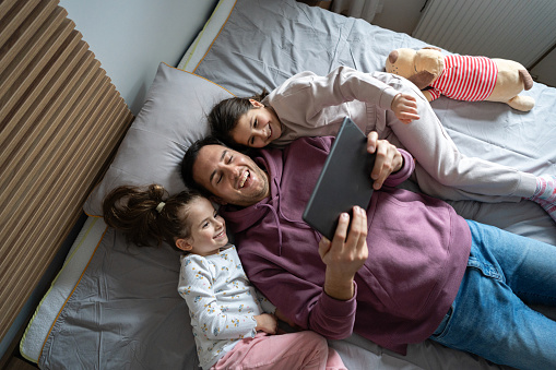 High angle of an young Caucasian father watching movie or cartoon on digital tablet while lying in the bed with his daughters