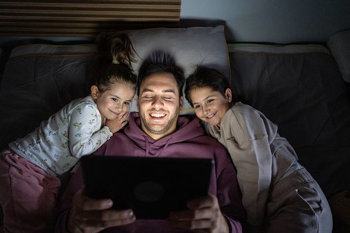 High angle of an young Caucasian father watching movie or cartoon on digital tablet while lying in the bed with his daughters