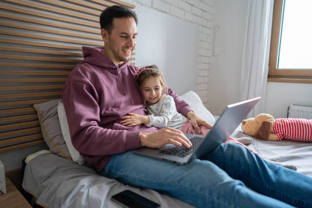Young father working on laptop, while lying in the bed with his daughter