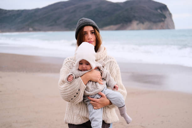 Mom holding her cute baby next to a sea wearing warm sweaters
