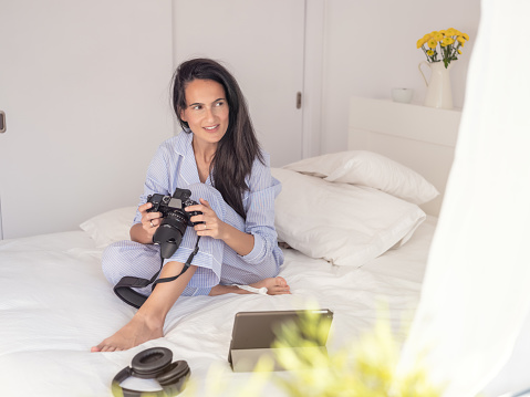 Dark haired woman with photo camera smiling and looking away while sitting on bed with gadgets in sunny morning