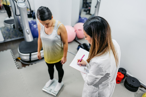 Young woman measuring her weight in the gym with her female nutritionist.