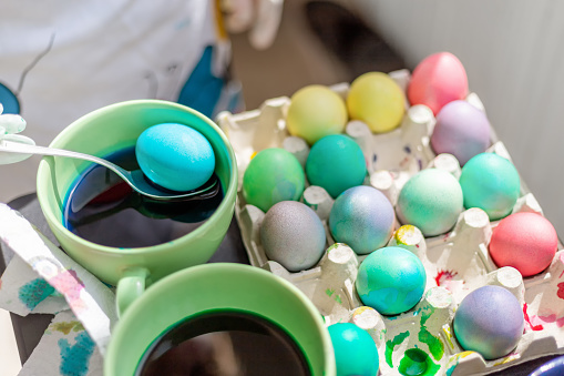 Coloring vibrant Easter eggs with kids at home, hand is dipping spoon and egg in blue liquid, messy table and color splash