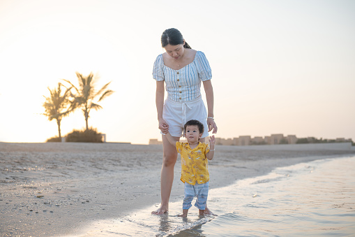 Caring young mother encourages her adorable baby boy to take his very first steps on the soft sands of the beach, while the waves wet their bare feet. Magical milestone captured in the golden glow of sunlight