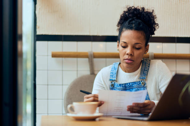 istock Serious-looking curly-haired female, holding one document, working at the cafe, using a laptop. 1962590134