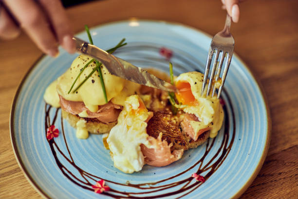 Close-up of female hands using a fork and knife, eating poached eggs with smoked salmon on toast.