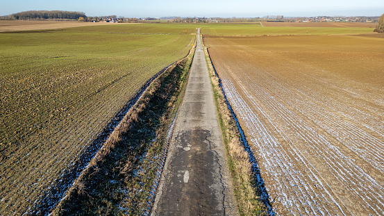 This image taken from above showcases a rural country road stretching into the distance, a straight line cutting through the varied tapestry of winter fields. On the left, the field shows signs of recent activity, with furrows and residual greenery hinting at a past harvest or preparations for spring planting. To the right, the field is covered with a dusting of snow, reflecting the season's chill and dormancy. The dividing road is bordered by hedgerows and occasional snow patches, evoking the solitude and quiet of a winter's day in the countryside. The distant horizon hints at the expanse of the agricultural landscape, a serene and enduring testament to the rhythms of nature and farming life. Seasonal Shift: Country Road Flanked by Winter Fields. High quality photo
