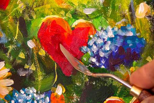 Hand of artist with palette knife close-up paints beautiful oil painting of red heart and flowers on canvas. Card for wedding and Valentine Day.