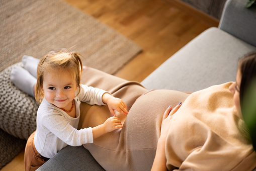 Happy little girl touching her pregnant mother's belly while her lying on sofa at home