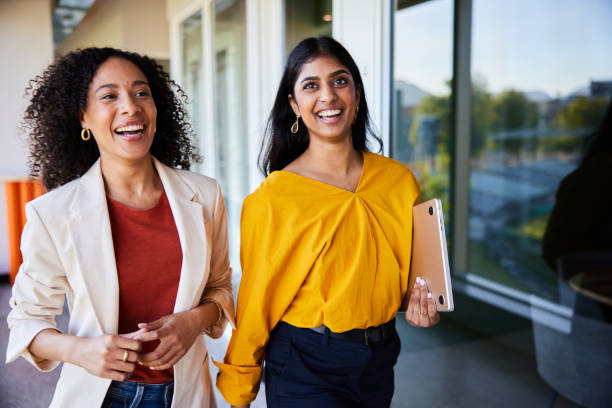 Diverse young businesswomen laughing together while walking in an office Two diverse young businesswomen talking and laughing together while walking down a corridor in a bright modern office indian woman smiling stock pictures, royalty-free photos & images