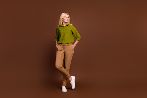 Full size photo of good mood woman dressed khaki shirt arms in pockets look at promo empty space isolated on dark brown color background.