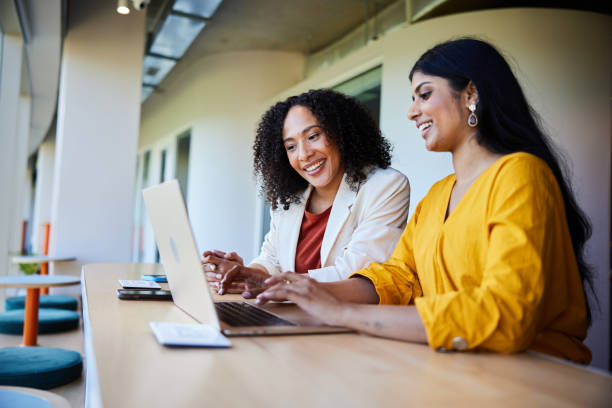 Two businesswomen smiling and working on a laptop at an office table Two diverse young businesswoman talking and working on a laptop together at a desk in an office lounge area indian woman smiling stock pictures, royalty-free photos & images