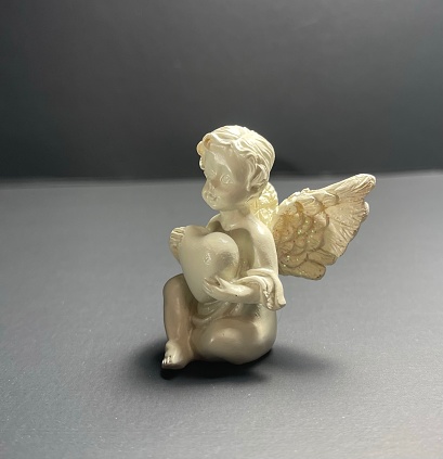 Porcelain angel figurine holding a heart, with detailed wings on a grey backdrop.