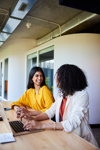 Two diverse young businesswoman talking and laughing while working on a laptop in an office lounge area