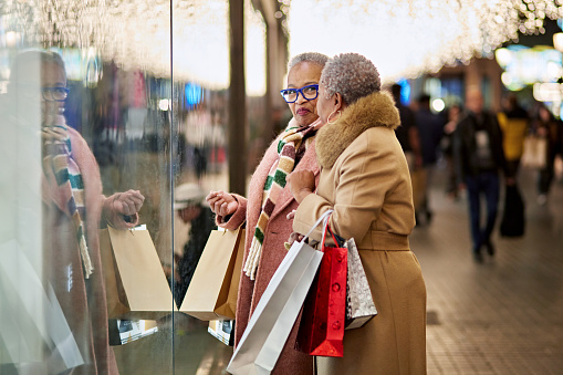 Side view of well dressed Black women in 50s and 60s holding bags, standing in front of retail window under glittering lights, face to face and smiling.