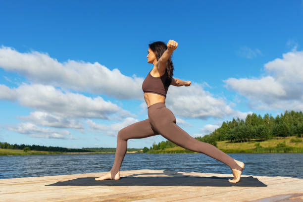 Attractive woman performing a variation of Virabhadrasana exercise, warrior pose, training in leggings and a short top while standing on the shore of a lake on a warm sunny morning stock photo