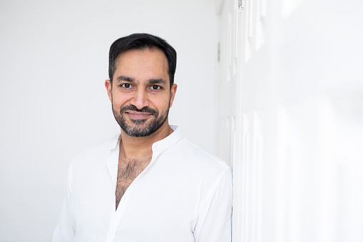 Relaxed portrait of a handsome man standing by white wardrobe doors in a bedroom. He is wearing a white shirt.