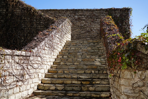 A stone staircase leading upwards, lined by a high stone wall on the right and a flat wall on the left. At the top you can see a blue sky with clouds