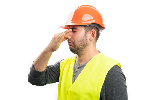 Builder man making grossed expression holding nose as bad smell concept using fingers wearing yellow fluorescent vest and orange helmet isolated on white background