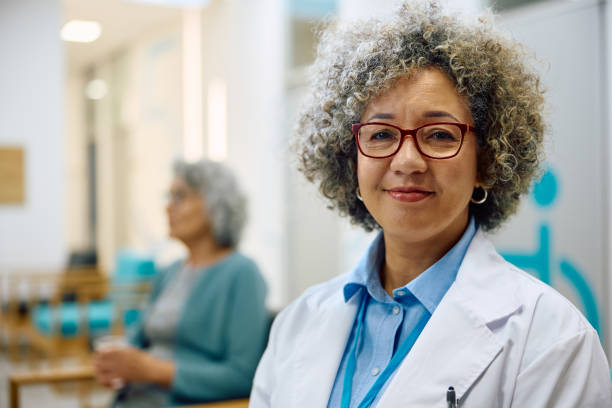 Portrait of smiling female doctor at medical clinic looking at camera. Smiling African American doctor in the hospital hallway looking at camera. Her patient is in the background. female doctor stock pictures, royalty-free photos & images