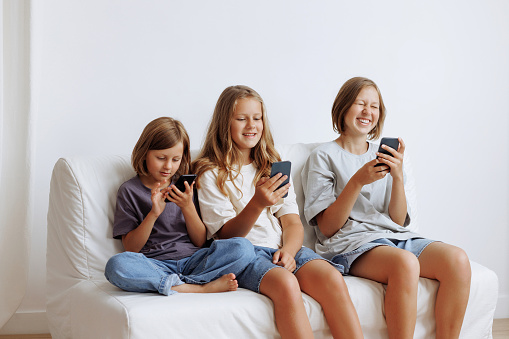Three girls use smartphones on the sofa. Three sisters smiling in casual clothes against a white wall. Concept for advertising a mobile application or content for the whole family.