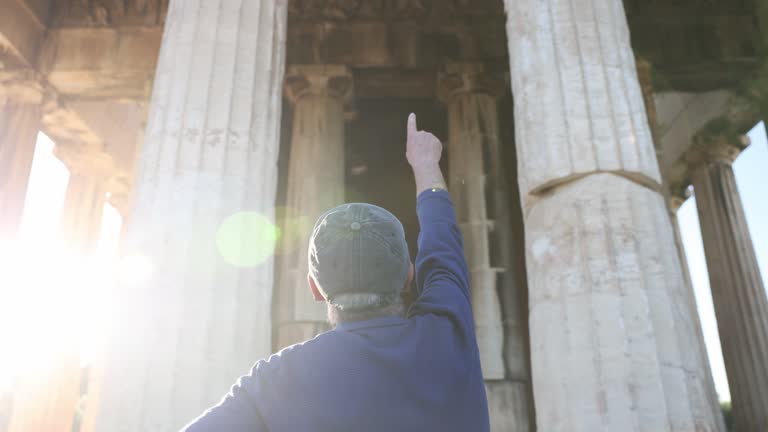 Adult man with a cap pointing to the Temple of Hephaestus, in the Ancient Agora of Athens (Greece) with a lens flare.