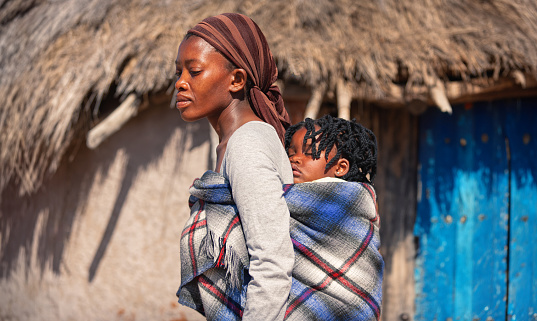 african mother in the village carry her child in the back in a traditional way in a blanket, background with the mud hut