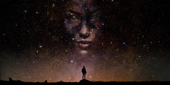 A silhouette of a person - either male or female, child or adult, standing on a rock outdoors, looking up at a night sky to see a huge face made up from the starfield. With copyspace.
