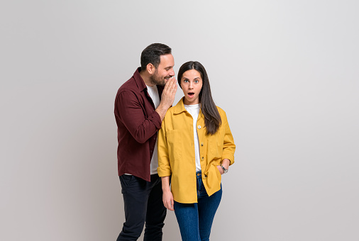 Young boyfriend whispering secret in shocked girlfriend's ear while standing on white background