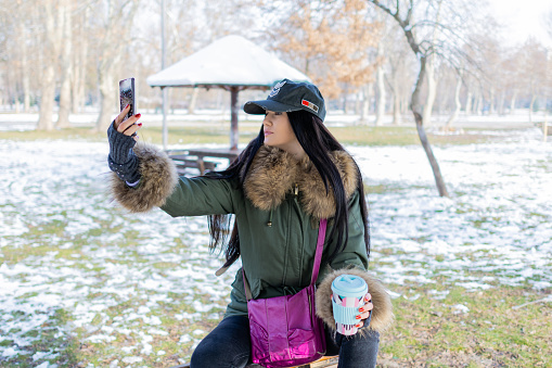 Beautiful young woman is enjoying her cup of coffee in public park on a snowy winter day,  Coffee break. She is taking selfie, or video call.